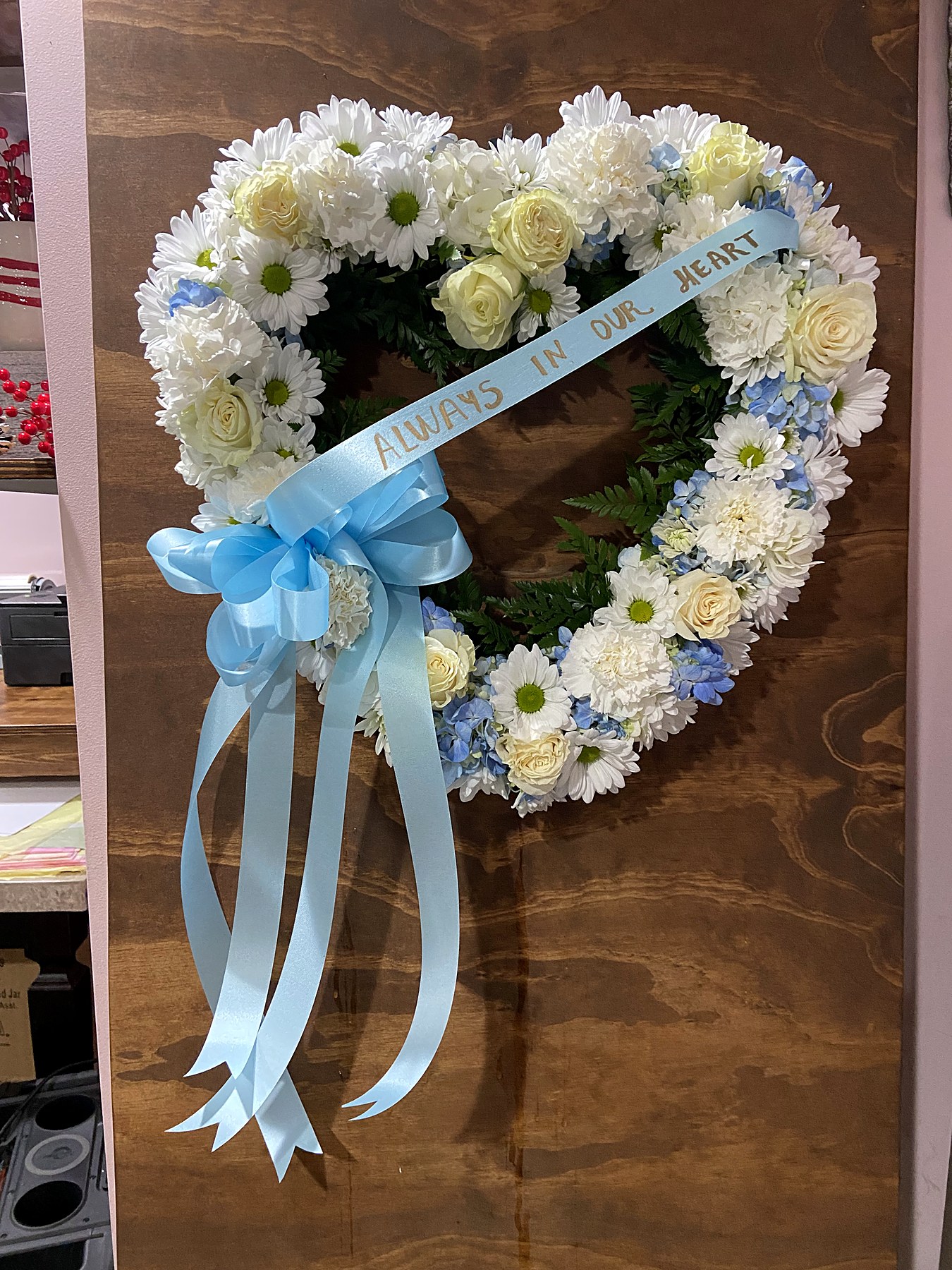 Heart-shaped tribute of white roses, daisies, and chrysanthemums with blue hydrangea and a blue ribbon